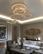 A tiered stack of lit gold rings hangs under a cove-lit ceiling above a white sofa and grey coffee table, shown as a Clérachic ring pendant.