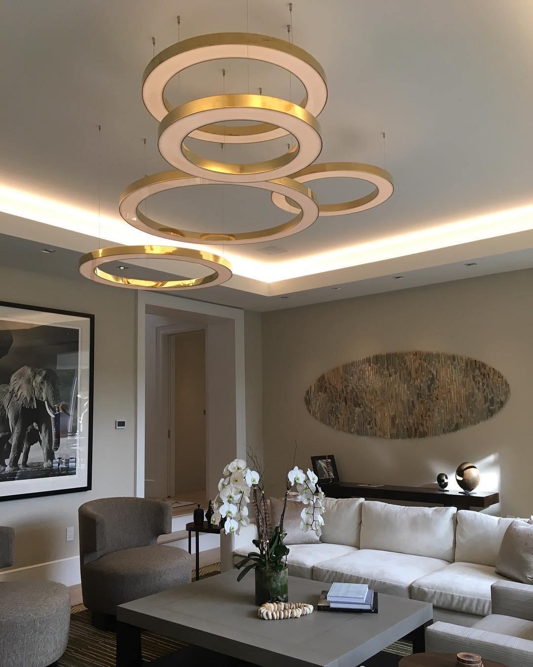 A tiered stack of lit gold rings hangs under a cove-lit ceiling above a white sofa and grey coffee table, shown as a Clérachic ring pendant.