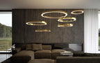 Gold rings hang at staggered heights over a dark lounge with a stone-clad wall and a window onto trees, lit by this Clérachic pendant.