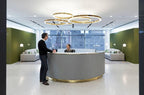 A cluster of gold rings hangs above a curved reception desk with a gold base, two people standing at it, lit by this Clérachic ring pendant.