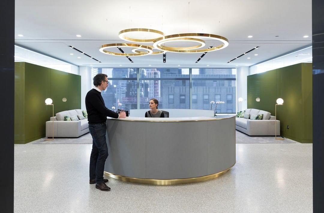 A cluster of gold rings hangs above a curved reception desk with a gold base, two people standing at it, lit by this Clérachic ring pendant.