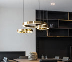 Three polished gold rings hang low over a dining table beside a black shelving unit with brass edges, lit by this Clérachic ring pendant.