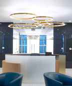 Gold rings hang at different heights above a curved white reception desk with blue armchairs, lit by this Clérachic ring pendant.