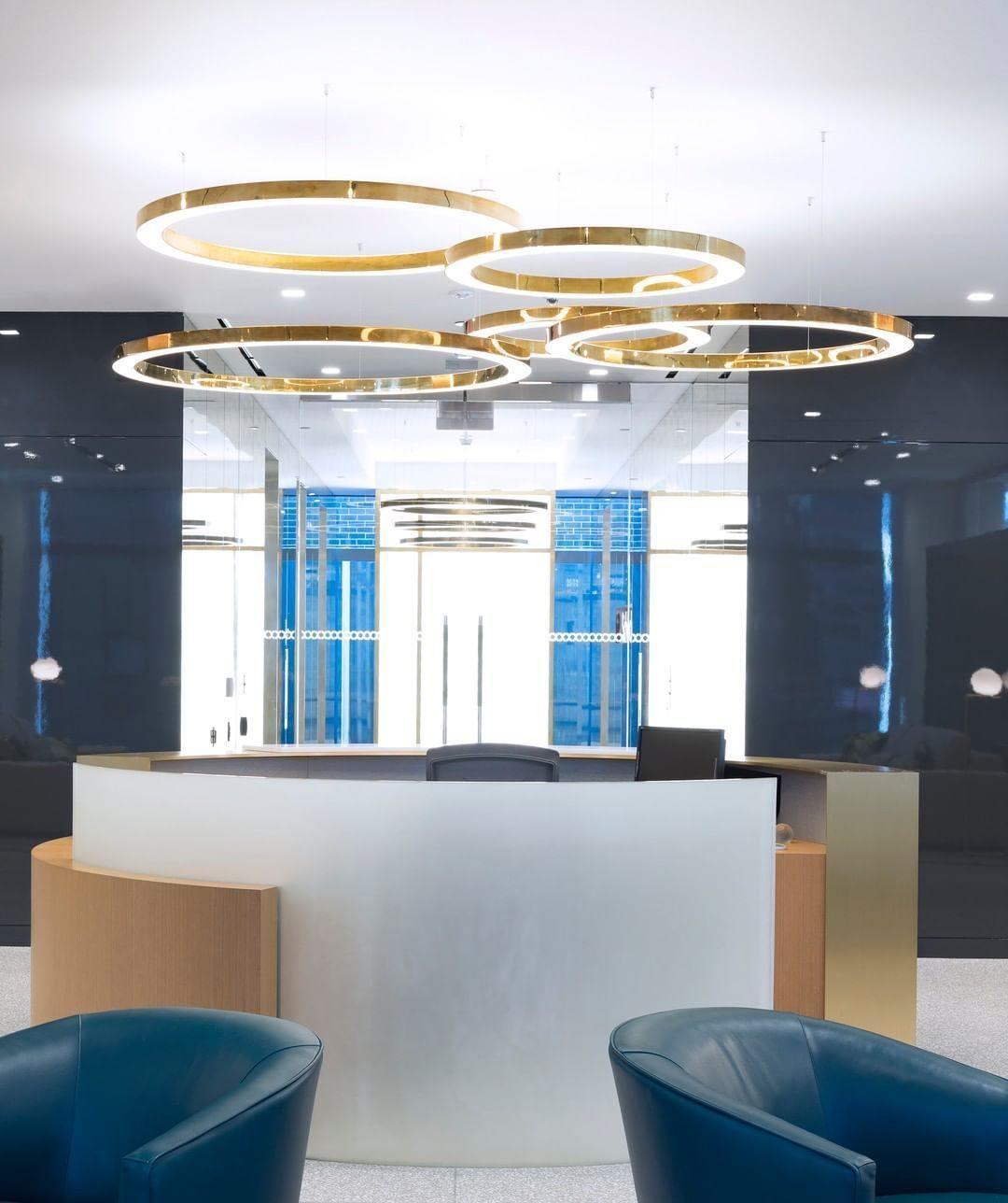 Gold rings hang at different heights above a curved white reception desk with blue armchairs, lit by this Clérachic ring pendant.