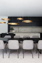 Five flat gold rings hang at varied heights above a long black dining table with grey upholstered chairs, lit by this Clérachic ring pendant.