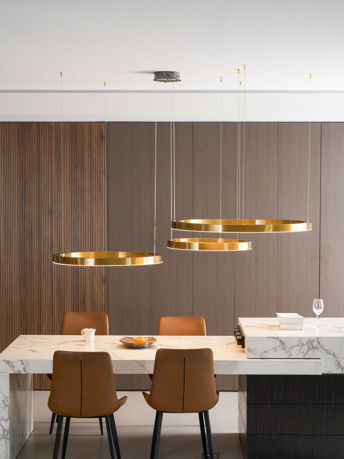 Three gold rings hang at different heights against a walnut-slat wall, above a marble counter and tan leather chairs, from Clérachic.