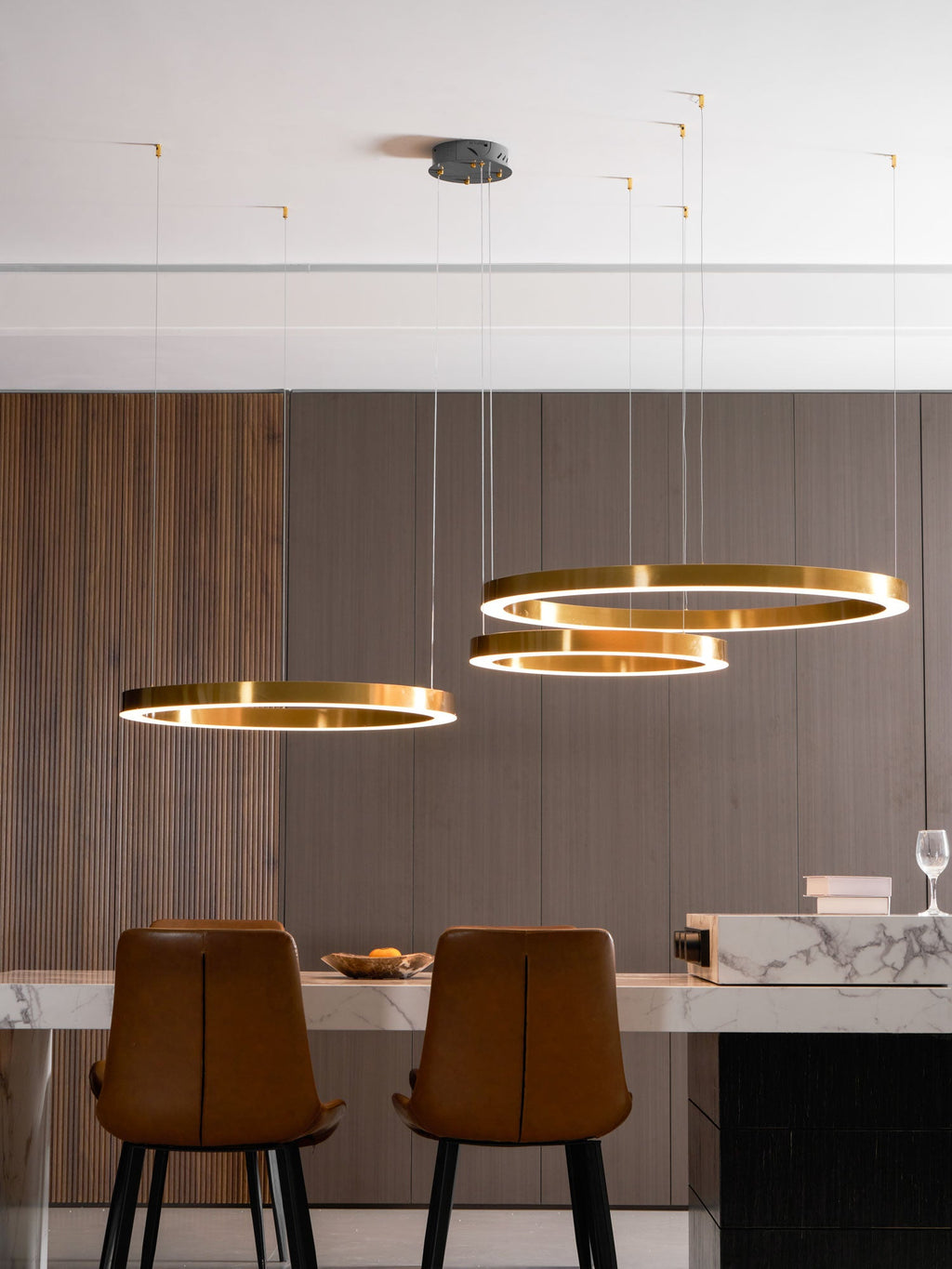 Three gold rings hang below a round ceiling plate, with tan leather chairs and a wine glass on a marble counter, lit by this Clérachic pendant.