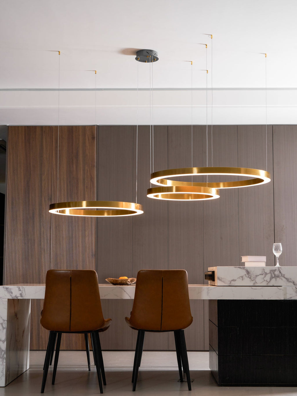 Three lit gold rings hang in a room with a walnut-slat wall, tan leather chairs and a white marble counter, from Clérachic.