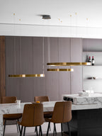 Three gold rings hang in front of a grey cabinet wall with bottles of red wine, above a marble counter, lit by this Clérachic ring pendant.