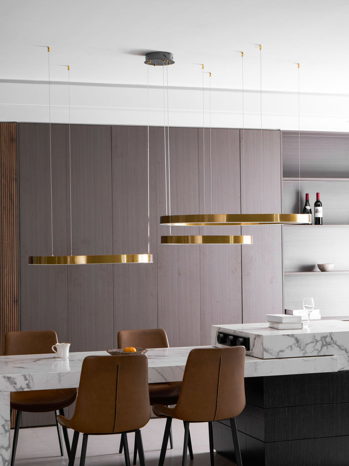 Three gold rings hang in front of a grey cabinet wall with bottles of red wine, above a marble counter, lit by this Clérachic ring pendant.