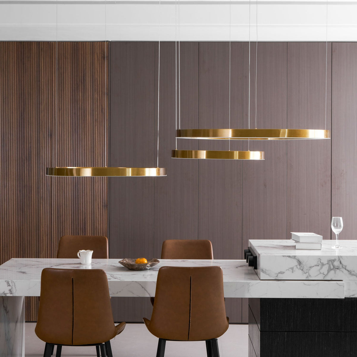 Three gold rings hang wide across the frame in front of a walnut wall above a marble counter with tan chairs, a Clérachic ring pendant.