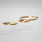 Three brushed gold rings hang at different heights against a soft grey studio wall, shown as this Clérachic ring pendant.