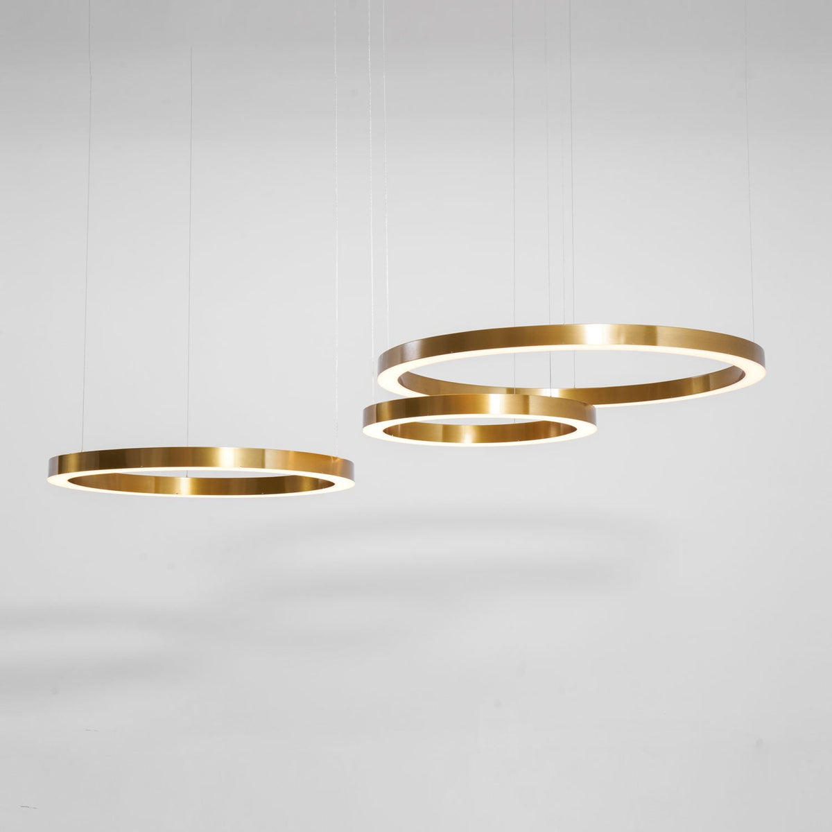 Three brushed gold rings hang at different heights against a soft grey studio wall, shown as this Clérachic ring pendant.