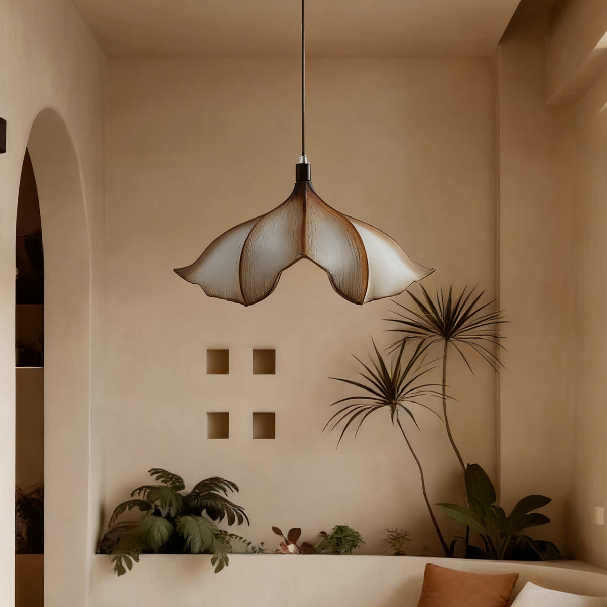 Unlit Clérachic petal pendant in front of a plaster wall with small square openings, palms and a curved bench below.