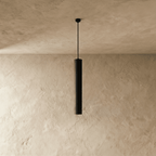 Clérachic slim black concrete cylinder pendant light, switched off, hanging from a thin black stem against textured plaster.