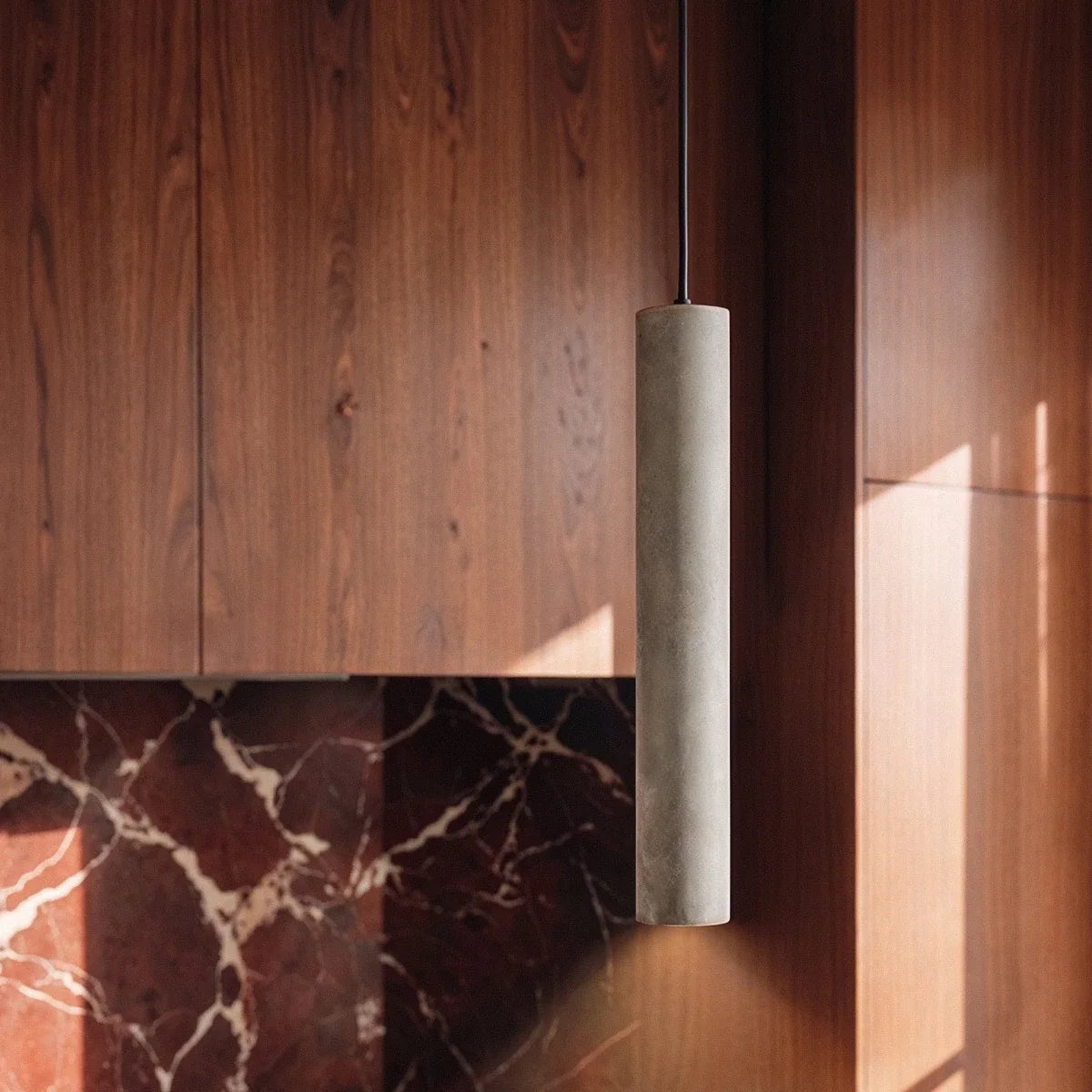 Clérachic close view of a slim grey concrete cylinder pendant light on a black cord beside walnut cabinets and red marble.