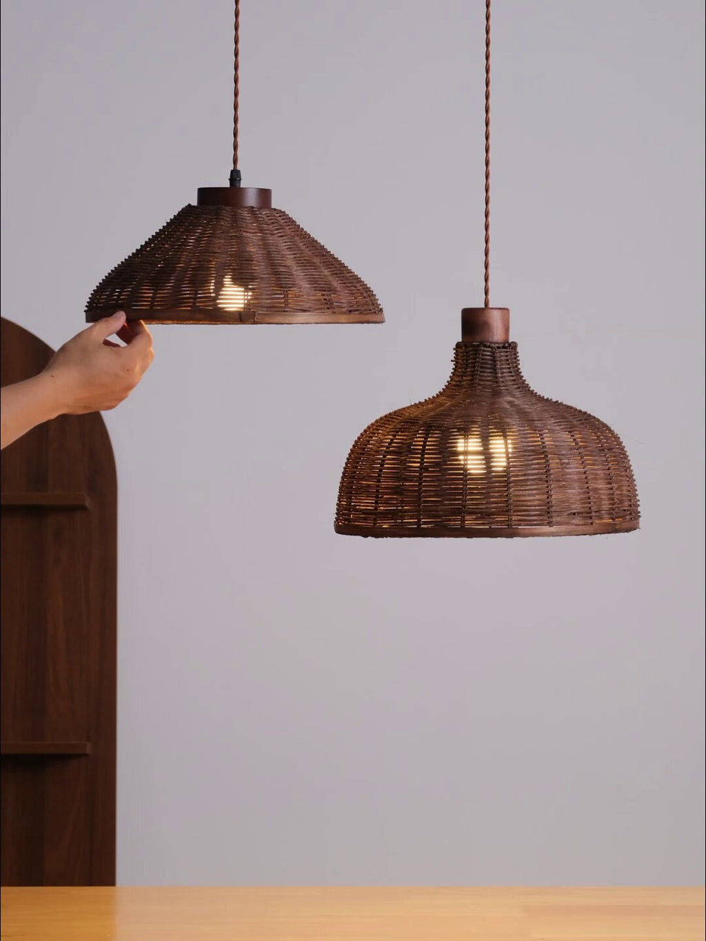 A hand reaches to adjust one of two rattan pendants from this Clérachic fixture, shown on a plain backdrop.