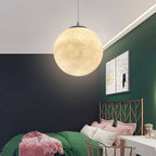 A single Clérachic globe pendant glows above a dark green and white wall, with a patterned print and a bed with pink cushions below.