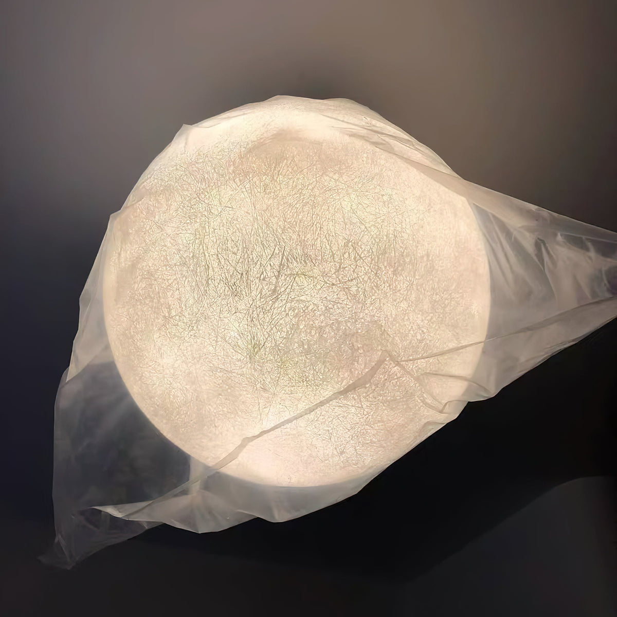 Looking up at a lit Clérachic sphere still wrapped in clear plastic, its fine, web-like fibre texture glowing through the film.