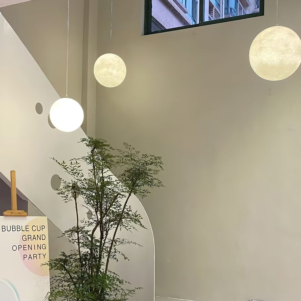 Three Clérachic spheres in different sizes hang at staggered heights in a tall stairwell beside a leafy indoor tree.