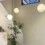 Three Clérachic spheres in different sizes hang at staggered heights in a tall stairwell beside a leafy indoor tree.