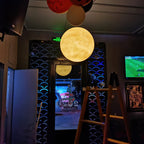 A warm yellow Clérachic sphere glows in a dark bar doorway, with a ladder and a screen showing a football match nearby.