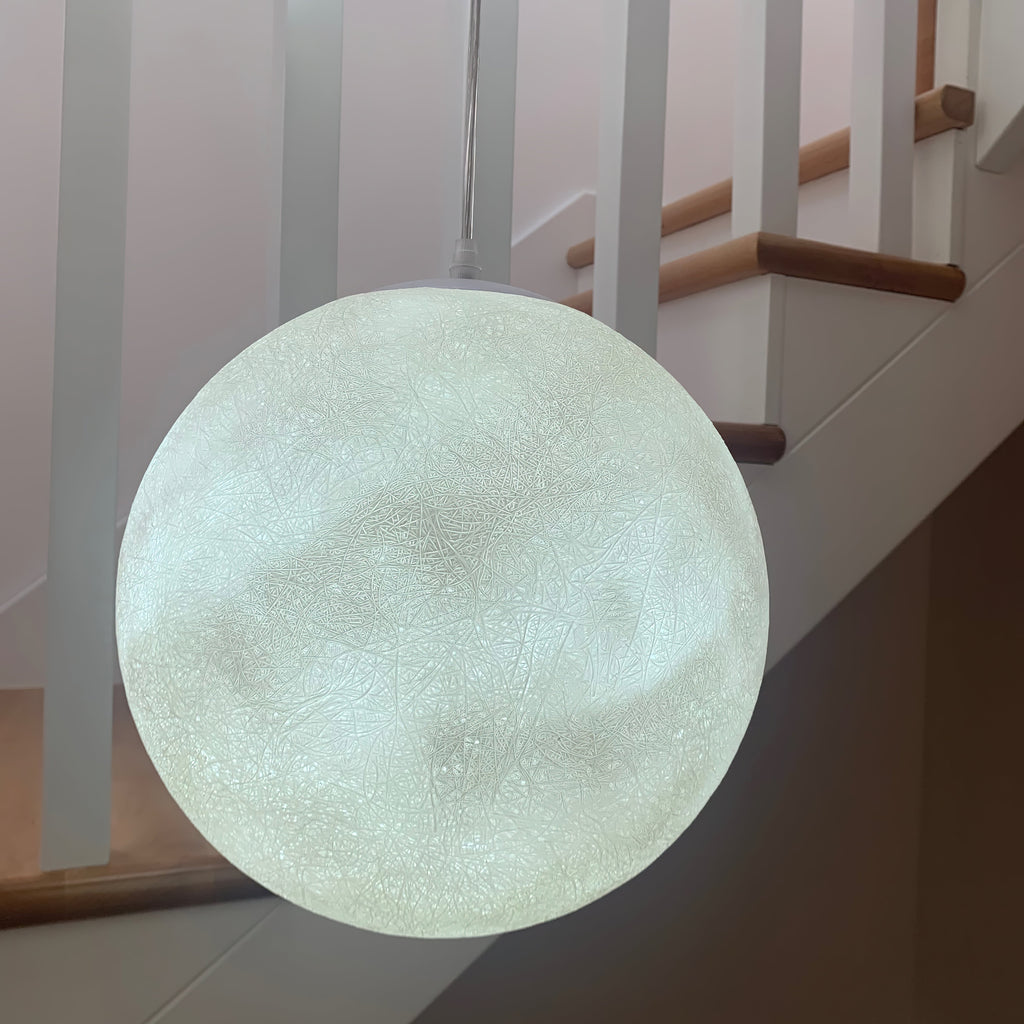 Close-up of a lit, pale green-white Clérachic sphere hanging in front of a white staircase and wooden handrail.