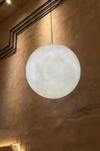 A lit white Clérachic sphere hangs against a rough brown plaster wall, with black conduit running across it.