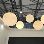 Large cream Clérachic spheres glow beneath a steel-trussed ceiling, with a dark wall and open space beneath.
