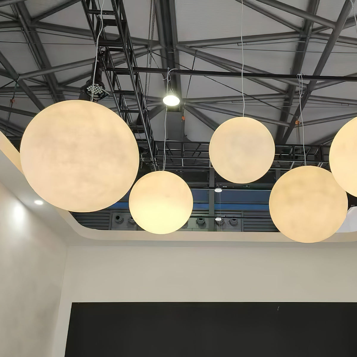 Large cream Clérachic spheres glow beneath a steel-trussed ceiling, with a dark wall and open space beneath.