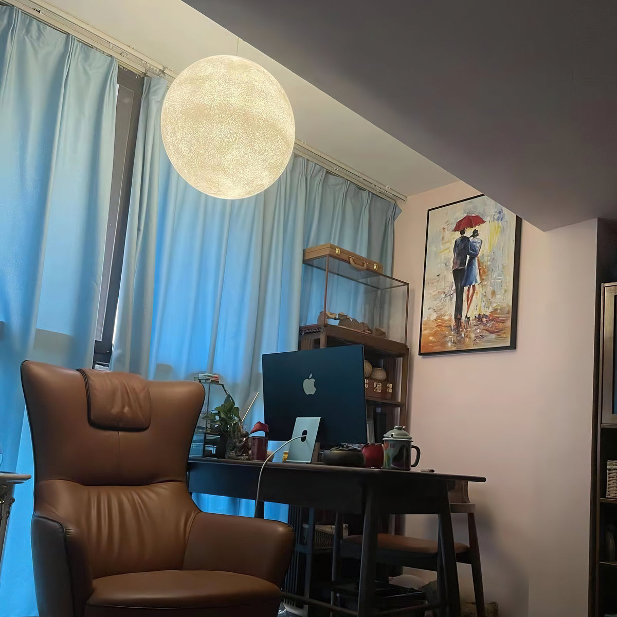 A pale Clérachic sphere hangs high beside a blue curtain, above a desk, a monitor and a tan leather armchair.