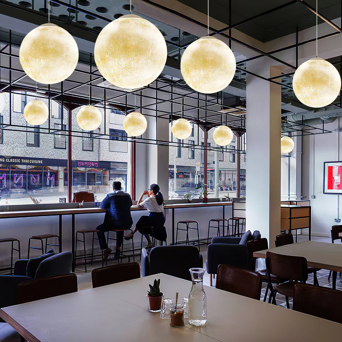 Cream Clérachic spheres of several sizes hang in a long row beneath a black steel grid, above a café table and window seats.