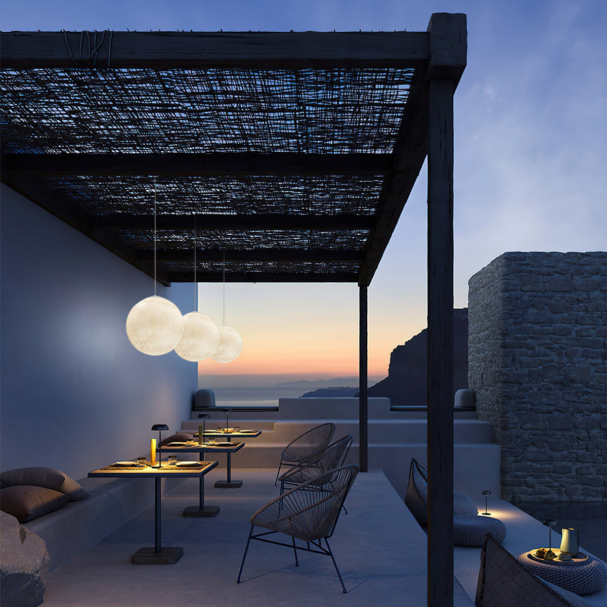 Three small Clérachic spheres hang under a dark timber pergola at dusk, looking over a terrace and a sea horizon.