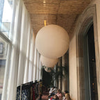 Two large matte peach-white spheres, a Clérachic-style pairing, hang in a corridor beneath a woven reed ceiling by tall windows.
