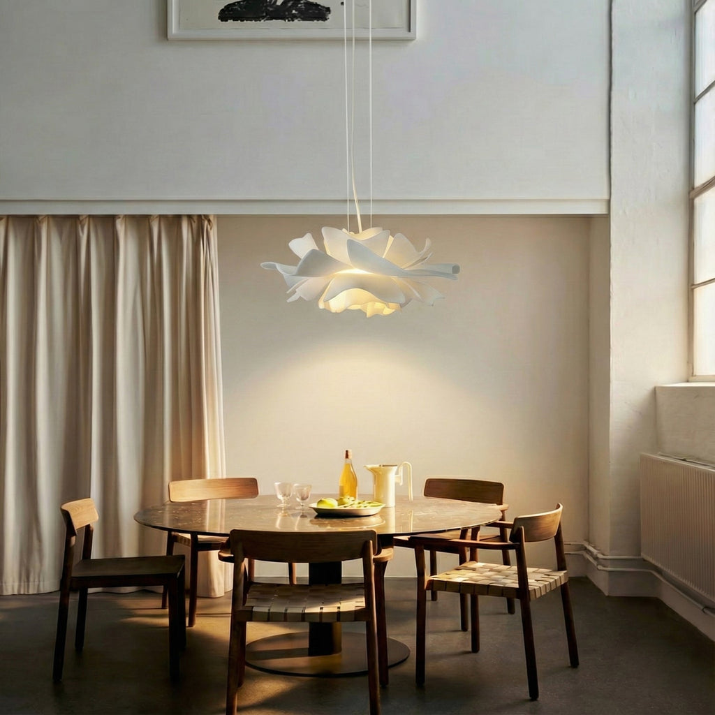 Lit Clérachic white petal pendant above a round dining table with dark-framed chairs, in a bright room with a tall window.
