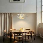 Lit Clérachic white petal pendant above a round dining table with dark-framed chairs, in a bright room with a tall window.