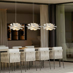 Three Clérachic white petal pendants in a row over a long table with white woven chairs, in a room with sheer curtains.