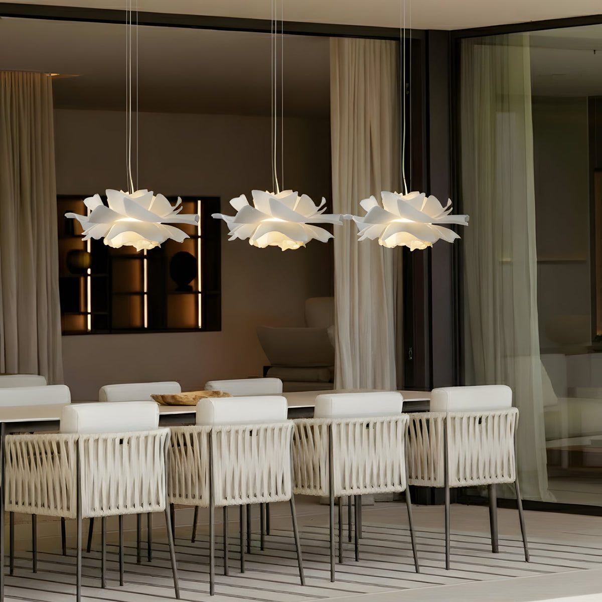Three Clérachic white petal pendants in a row over a long table with white woven chairs, in a room with sheer curtains.