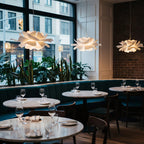Clérachic white petal pendants hung at different heights in a restaurant, above round tables and a blue-green banquette.
