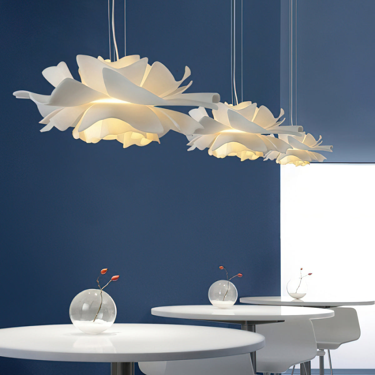 Three Clérachic white petal pendants above round white tables, against a deep blue wall with small glass vases.