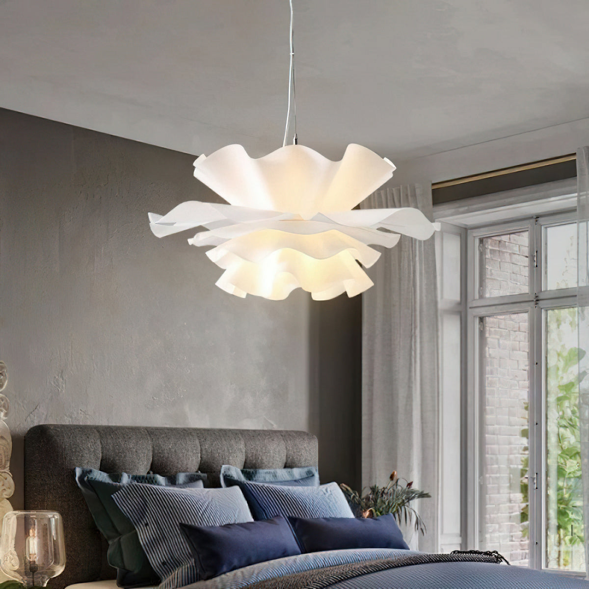 Large Clérachic white petal pendant glowing above a grey tufted bed with navy cushions, beside a tall window.