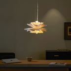 Lit Clérachic white petal pendant with a warm glow under the petals, above a desk with a mug and a closed laptop.