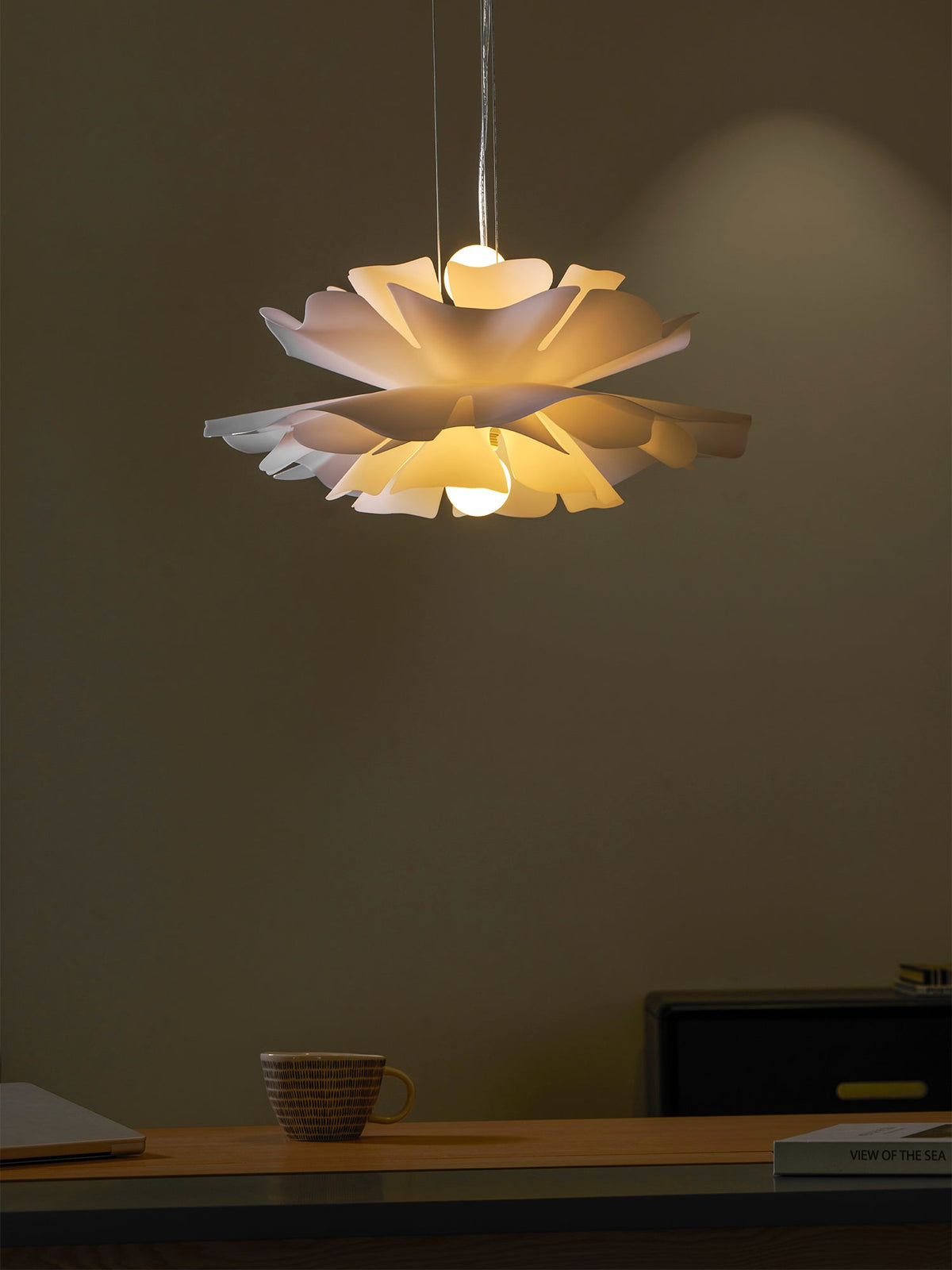 Clérachic white petal pendant lit warmly, with a bulb above and below the layers, over a desk and black cabinet.