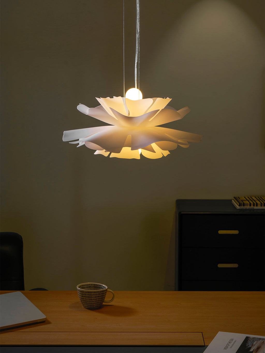 Clérachic white petal pendant glowing with a warm bulb at the centre, in front of a dim wall and a wooden desk.