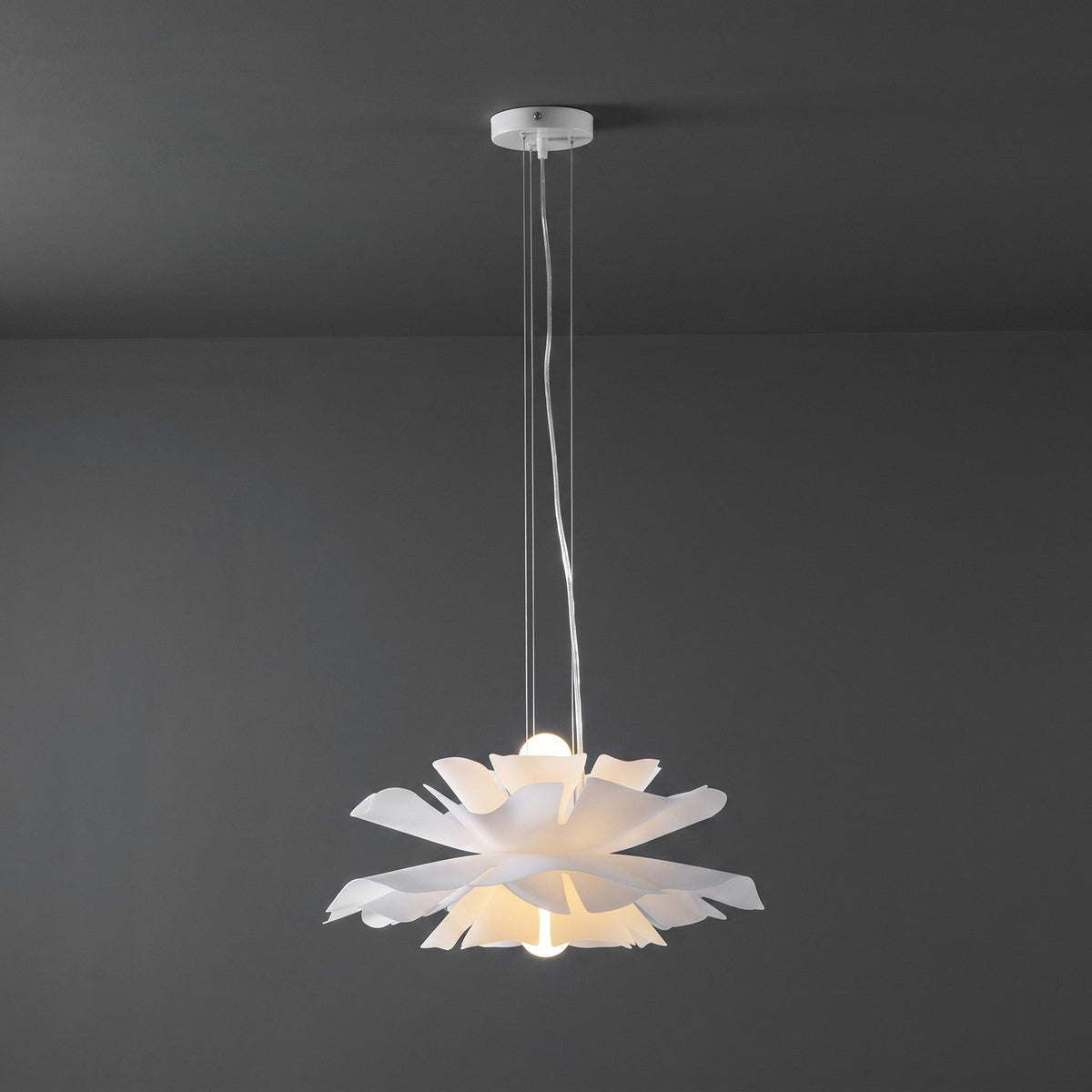 Clérachic white petal pendant with a white ceiling canopy, lit, with a bulb glowing inside the petals, on a dark grey wall.