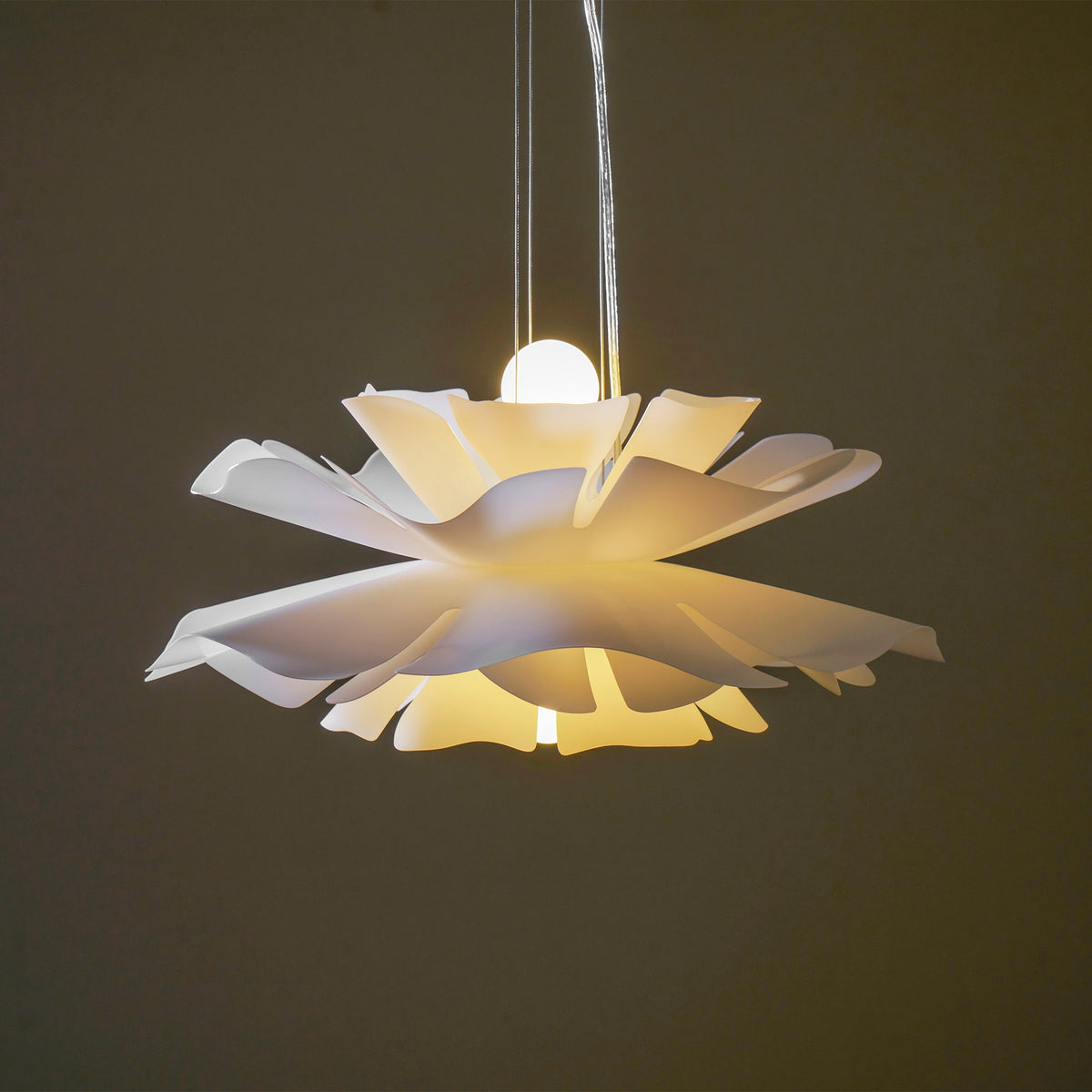 Lit Clérachic white petal pendant with a round bulb above and warm light spilling under the petals, on a dark olive backdrop.