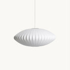 On a plain white backdrop, a wide, flat white silk pendant from Clérachic hangs from a thin cord, its ribbed shade glowing softly.