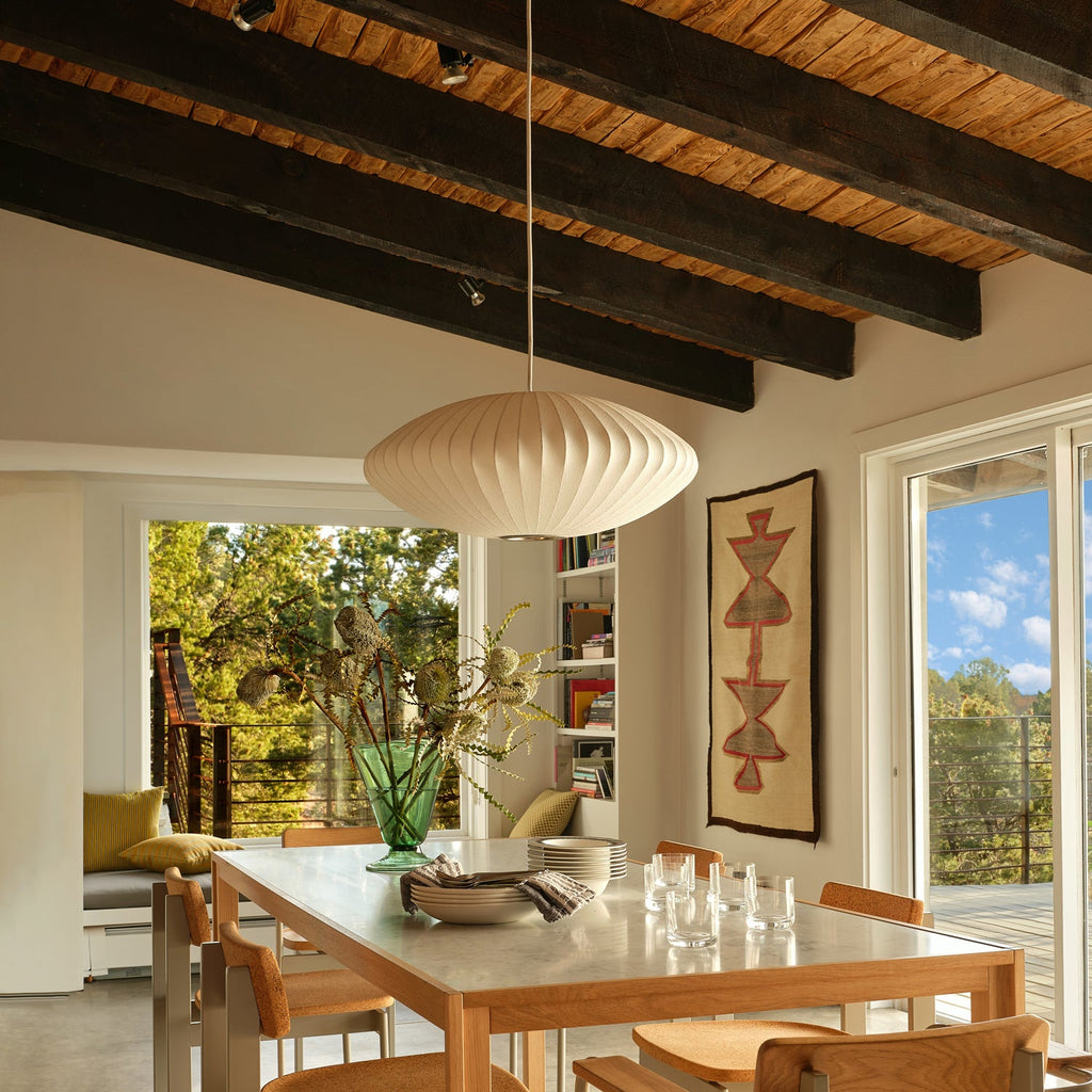 A Clérachic pendant with a white ribbed shade hangs above a long dining table, below a dark-beamed ceiling and beside tall glazed doors.