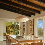 A Clérachic pendant with a white ribbed shade hangs above a long dining table, below a dark-beamed ceiling and beside tall glazed doors.