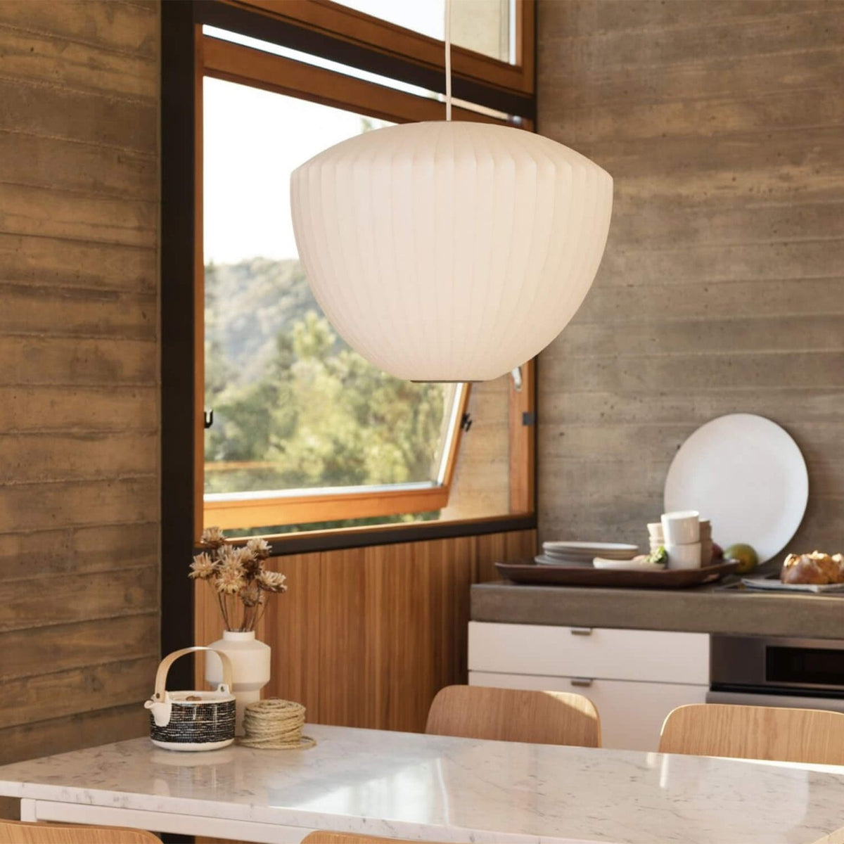 A rounded white Clérachic pendant with a softly ribbed shade hangs over a marble counter beside a wood-framed window.